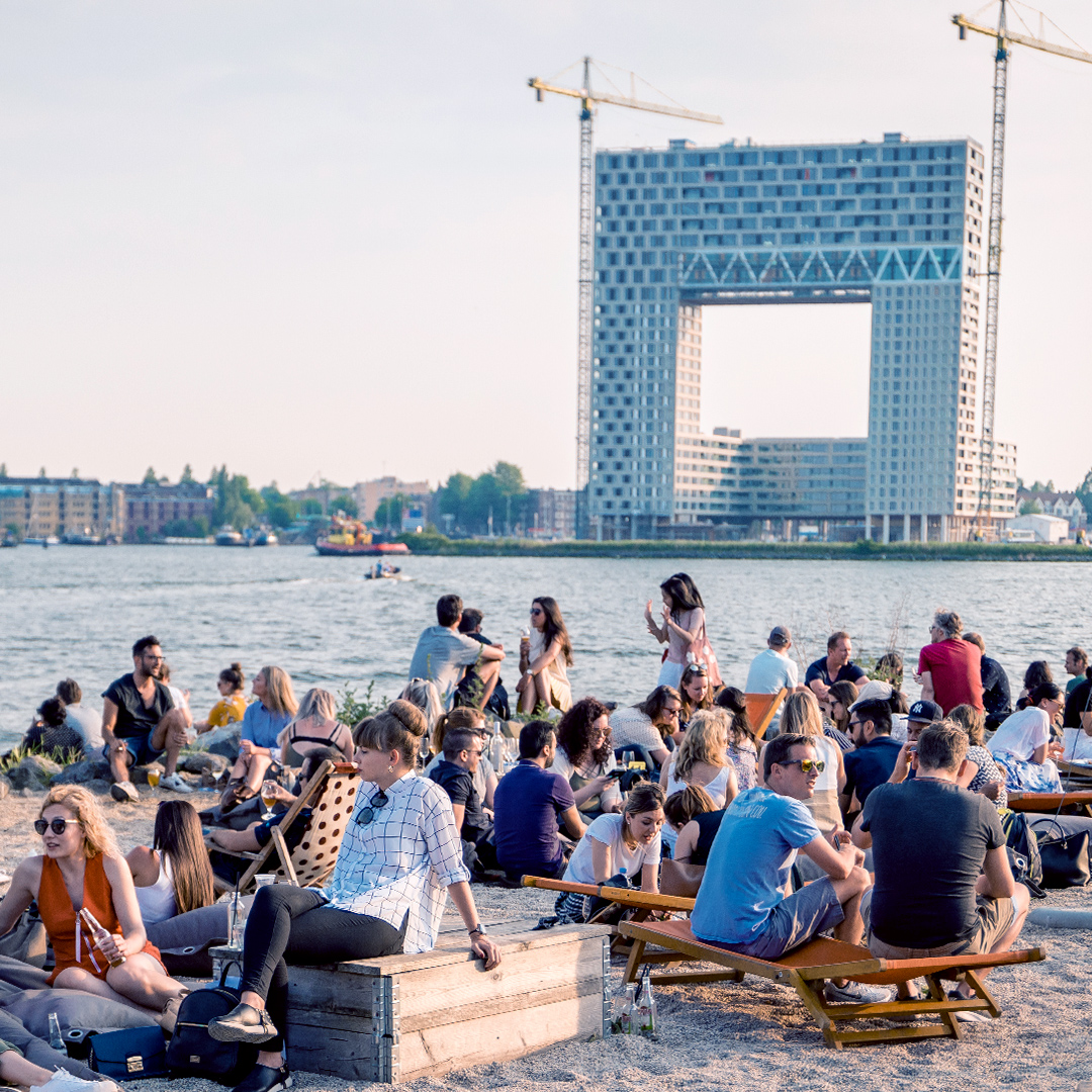 People sit on deckchairs on sand next to river with construction backdrop