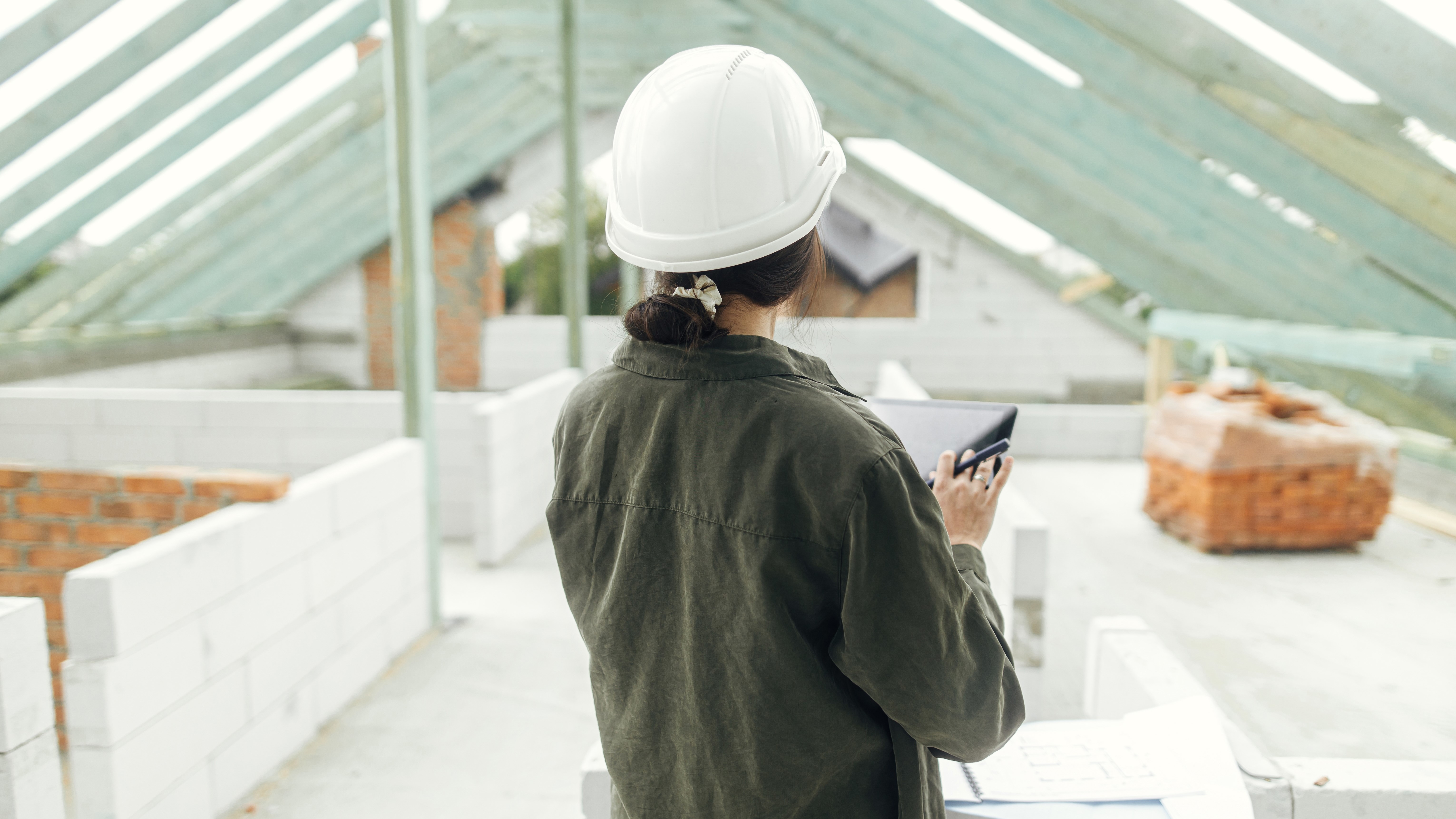 Young female architect with tablet checking blueprints against wooden roof framing of modern farmhouse. Stylish woman engineer in hard hat looking at digital plans at construction site; Shutterstock ID 2159615445; purchase_order: N/A; job: Sarah Hayford; client: RICS; other: 