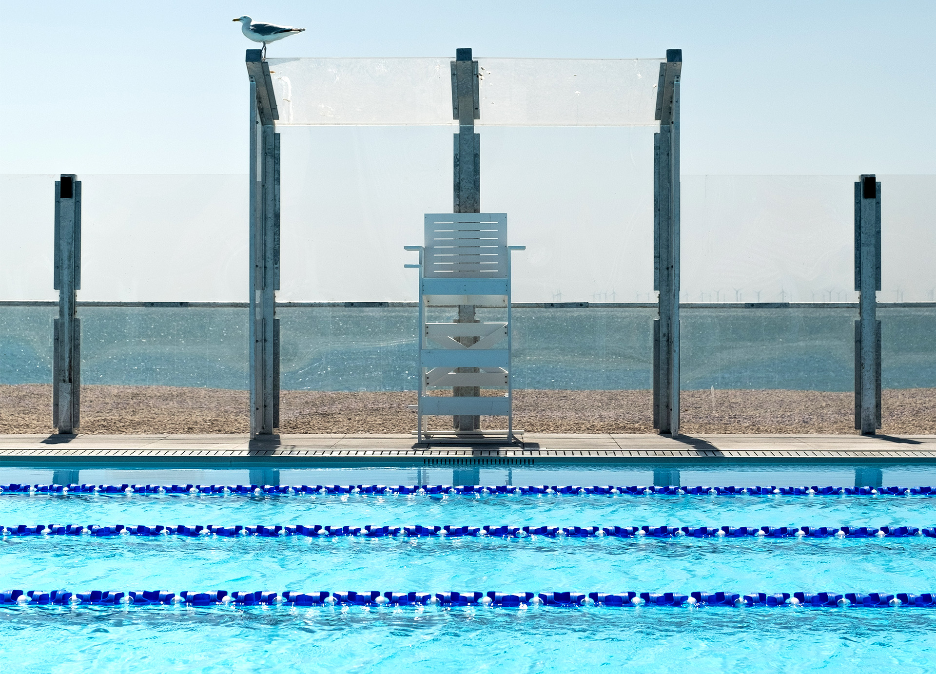 Lifeguard chair sits on the edge of outdoor pool, with the beach and sea behind. A seagull sits on top of the fence
