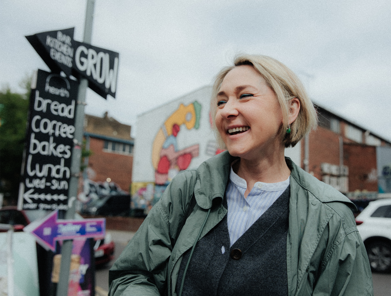 Yulia Pak MRICS portrait Yulia in a green raincoat and grey cardigan stands looking off camera in front of a graffitied warehouse and a sign post towards a cafe