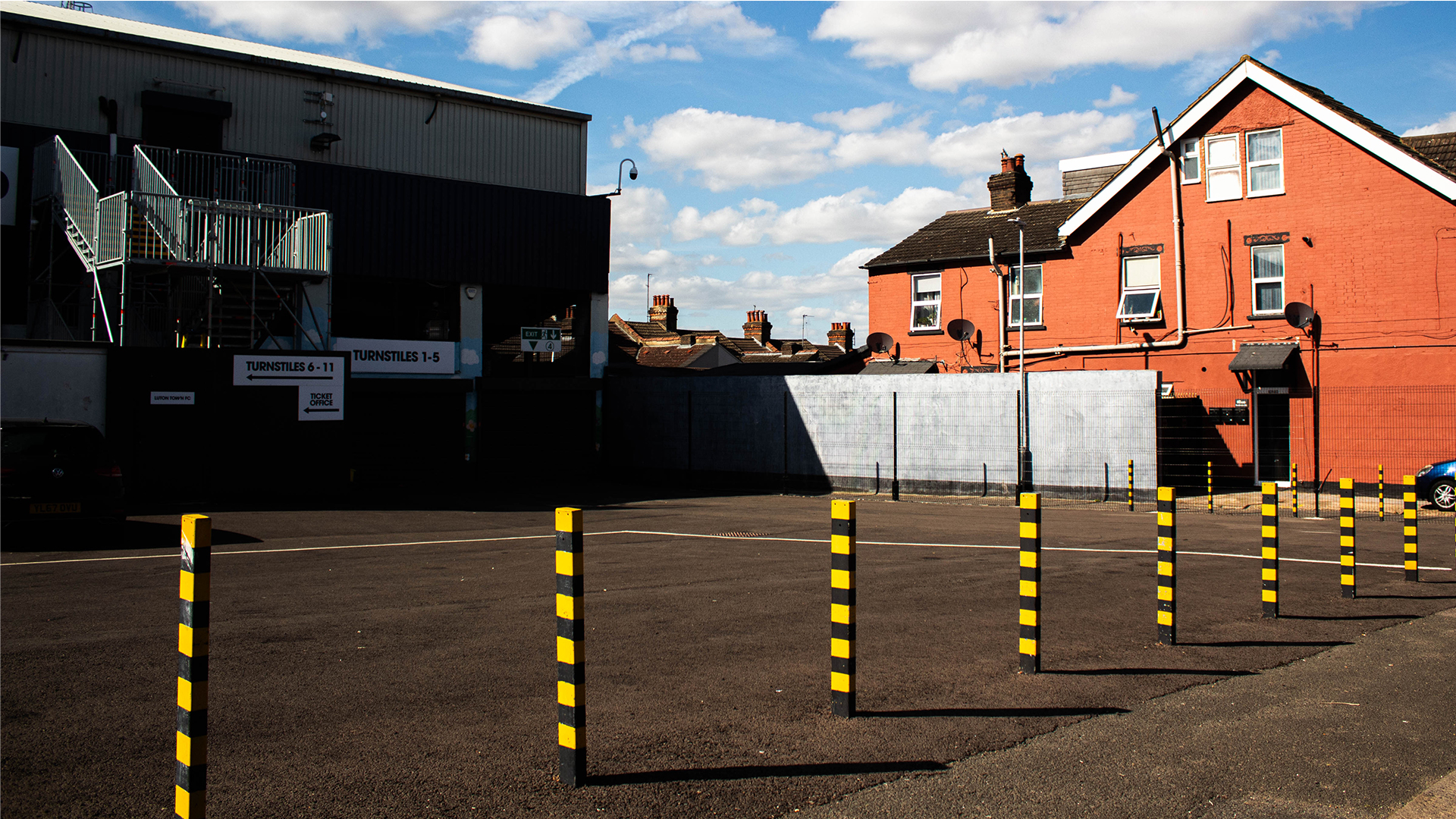 Empty car park in front of the stadium with yellow and black striped bollards and house in background