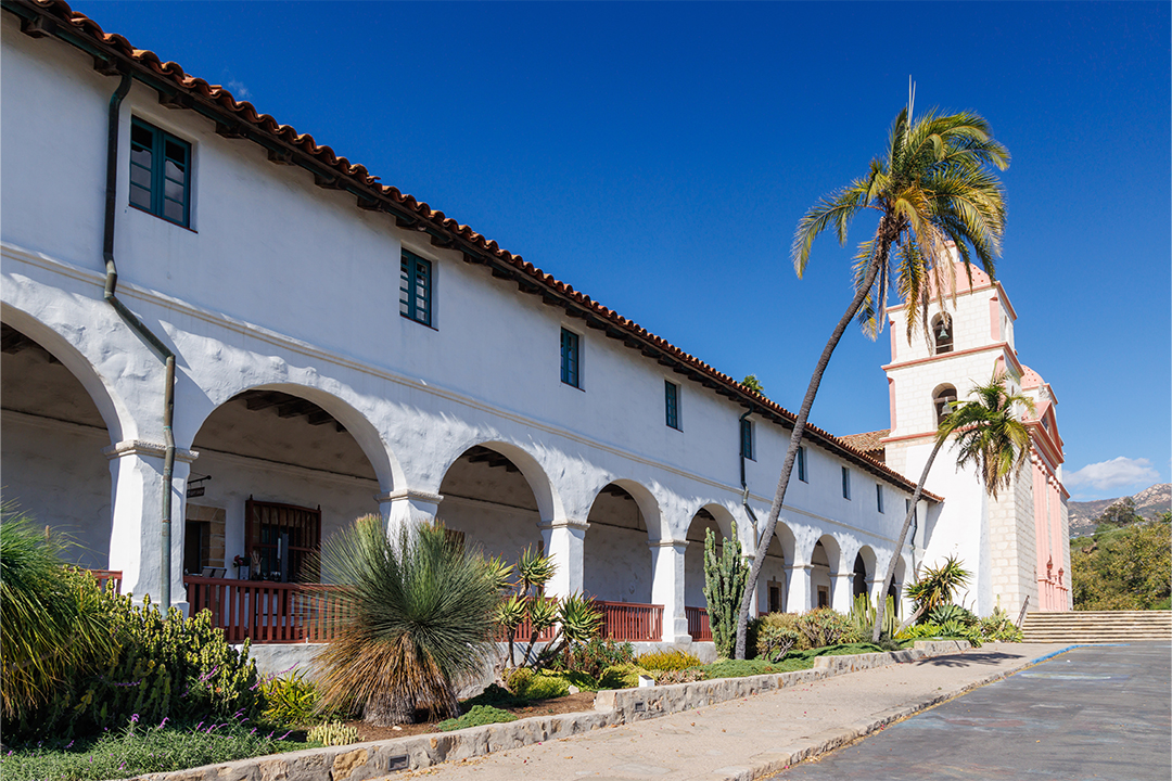 Queen of the Missions Church in Spanish colonial architecture style, with arched walkway, twin bell towers, and palm trees