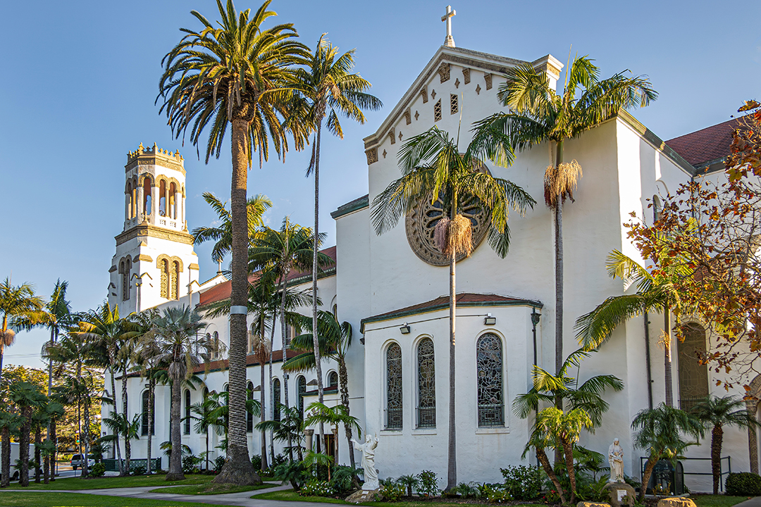Our Lady of Sorrows Church Beige west facade of Our Lady of Sorrows church with sunlight on tower against blue sky. Green palm tree foliage and lawn in front