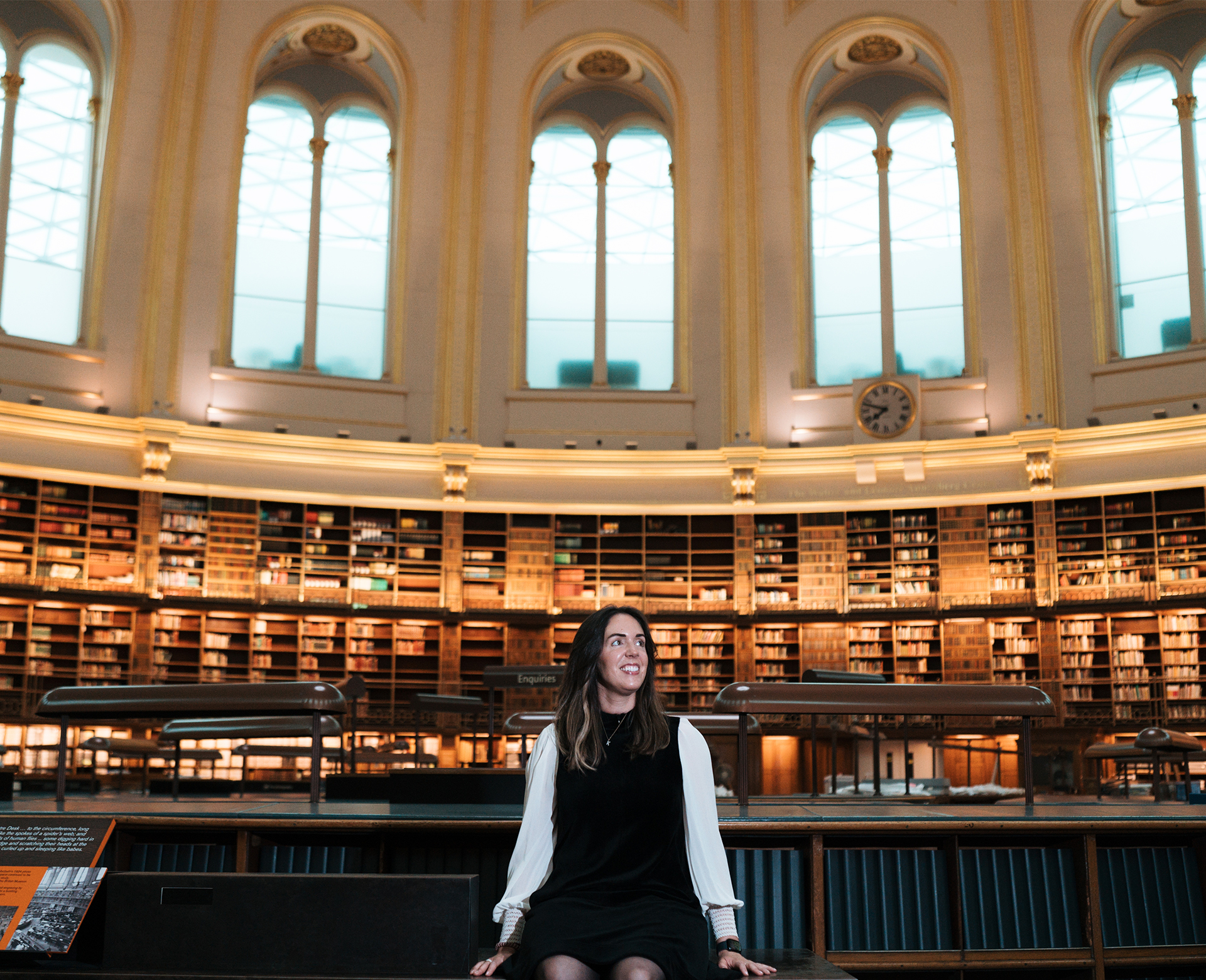 Katharine sits on a table surrounded by desks and shelves of books in a domed library