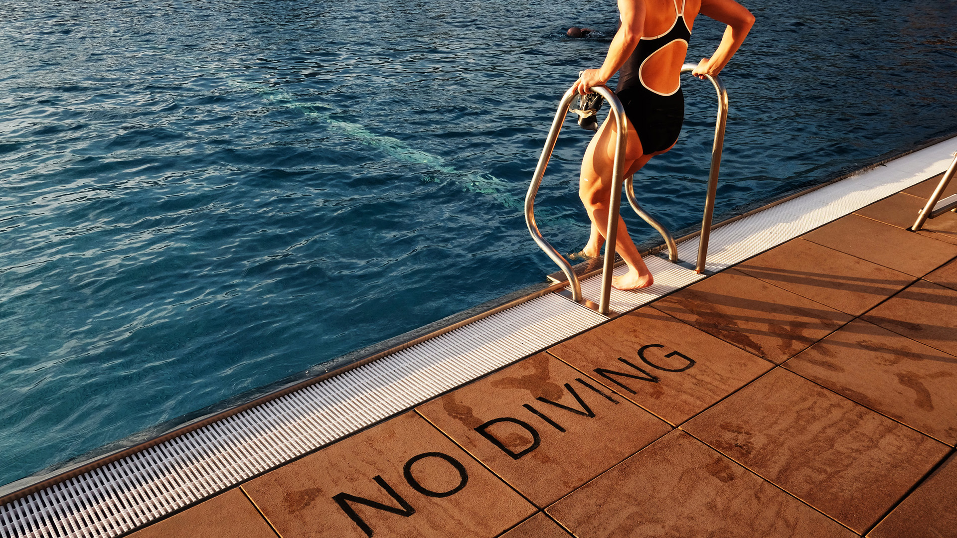 Swimmer walks down the stairs into an outdoor swimming pool with sign 'no diving'