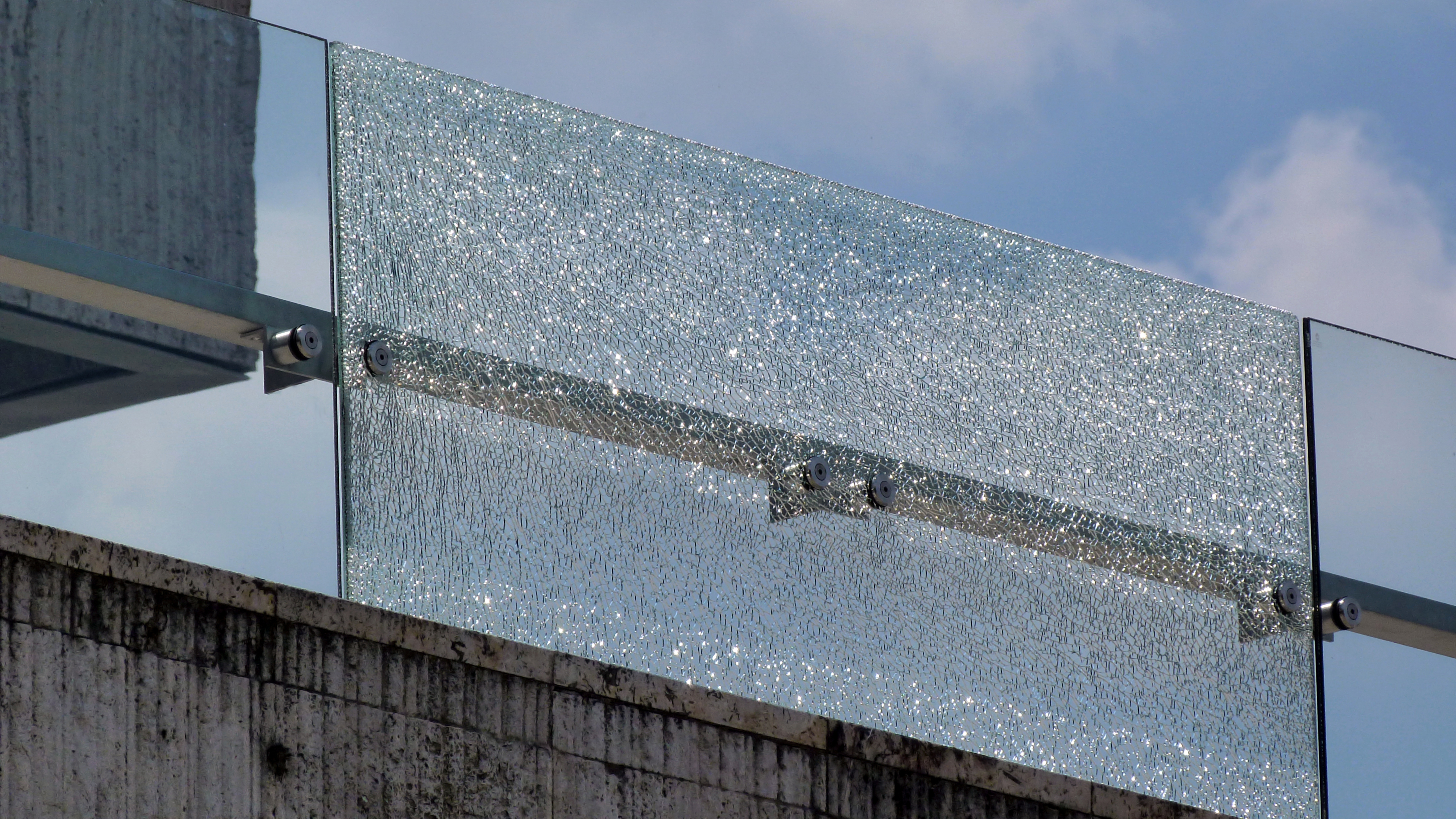 Image of shattered glass on a balcony panel on a concrete building, against blue sky