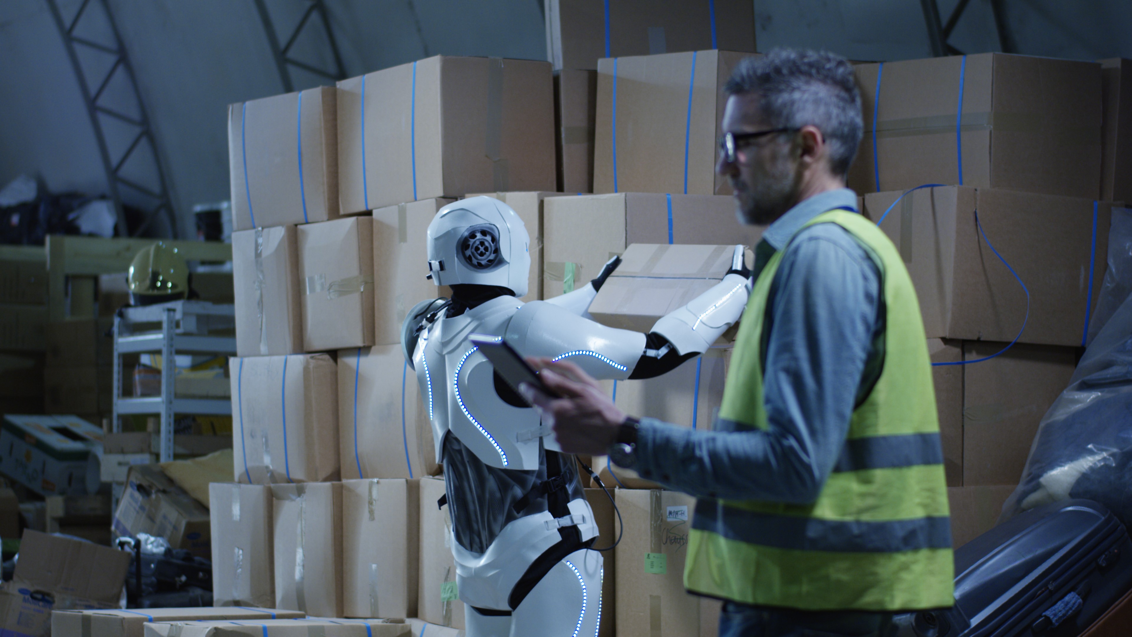 Man with digital pad alongside robot lifting cardboard box inside warehouse