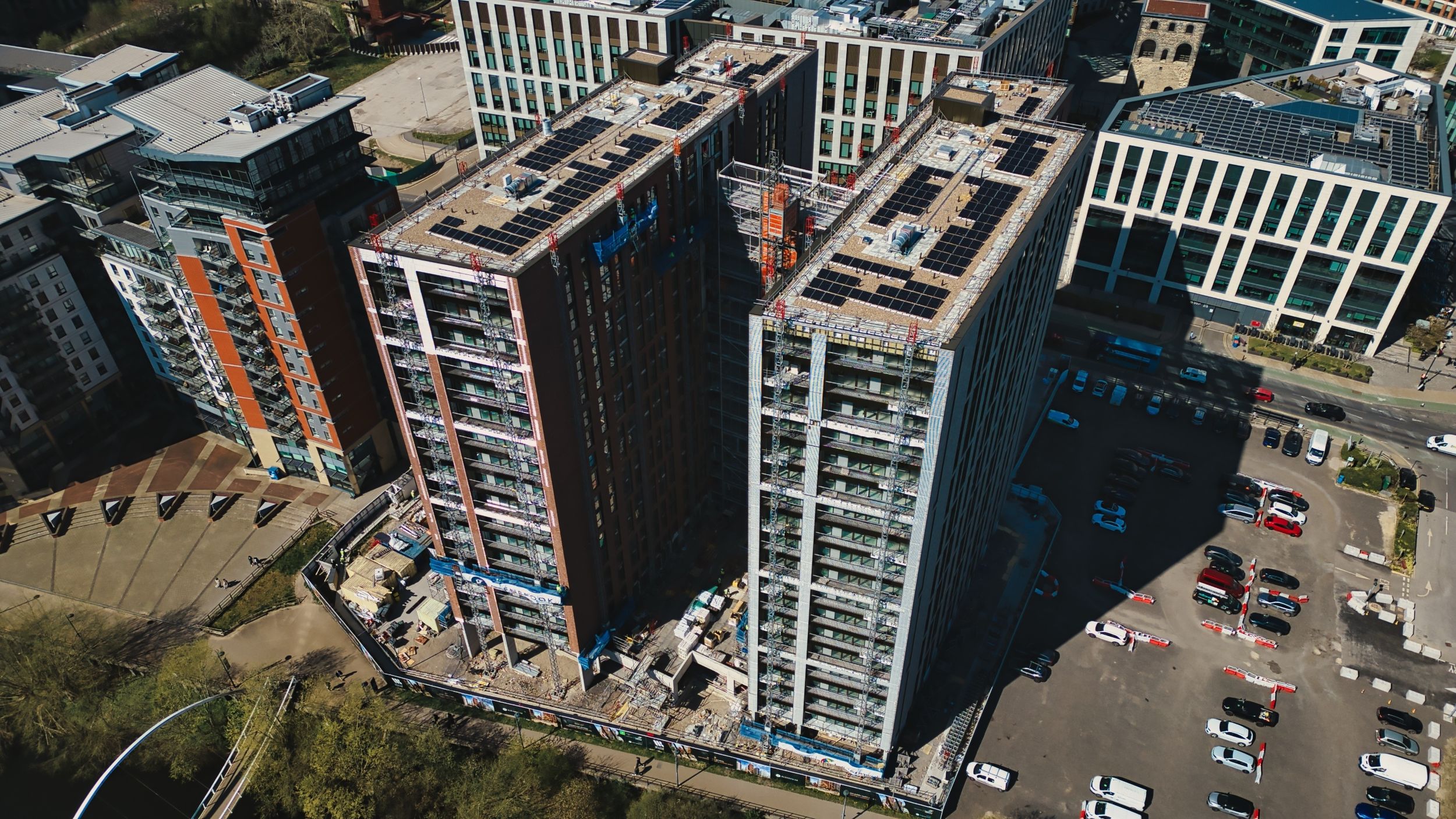 Aerial view shows high-rise buildings, some under construction, with parking lots and streets filled with cars in a modern urban environment in Leeds, UK.