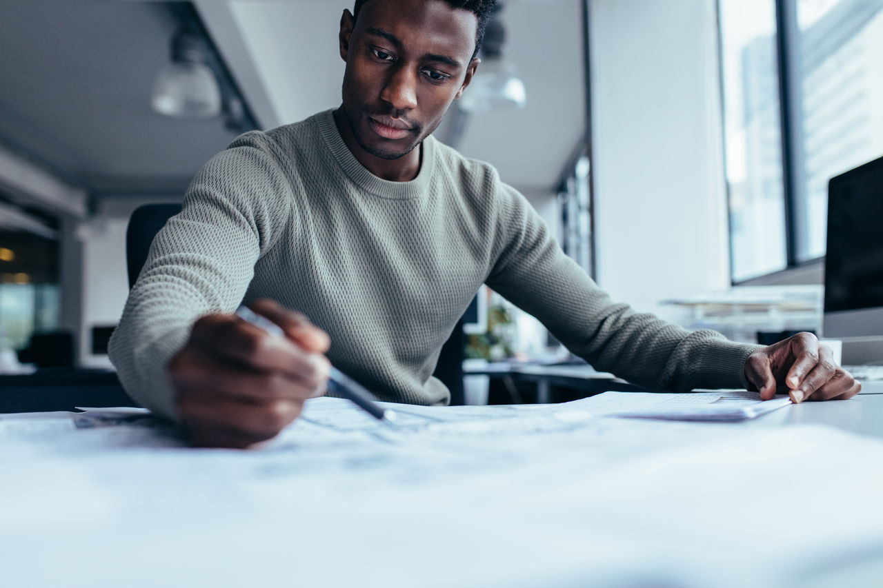 Businessman pointing at building plan in office. Black man working on construction plan.; Shutterstock ID 721778476; purchase_order: -; job: -; client: -; other: -