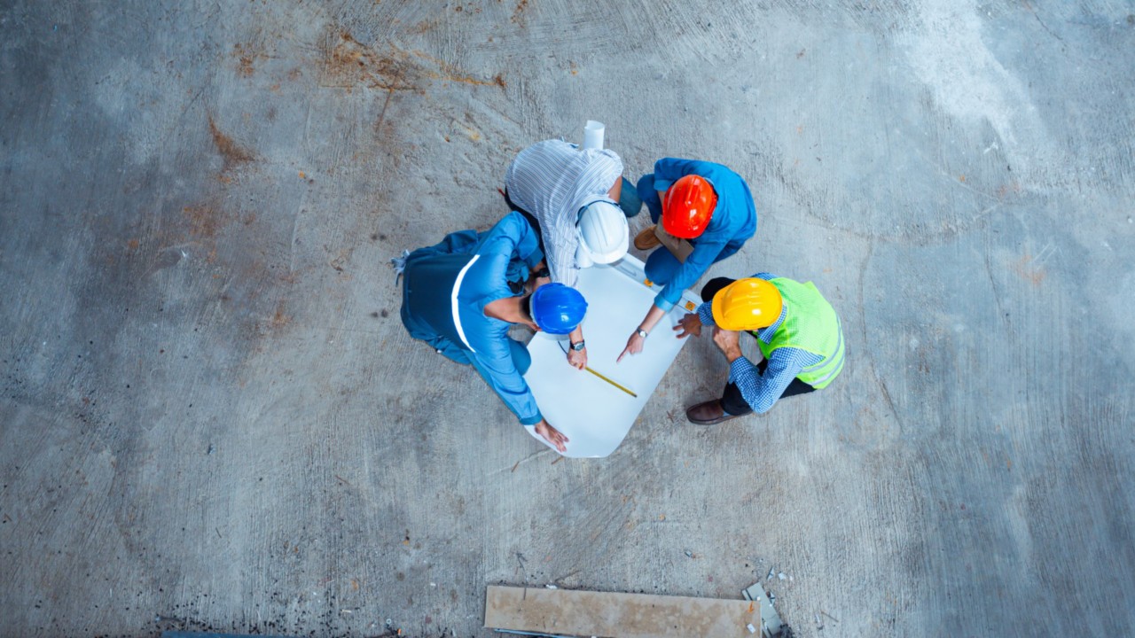 Four construction professionals with different coloured hats talk while leaning over a design board on a construction site